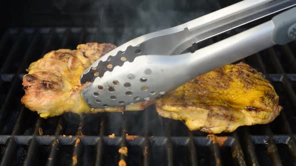 The Chef Turns A Piece Of Chicken On The Steaming Grill Of A Barbecue With Metal Tongs.