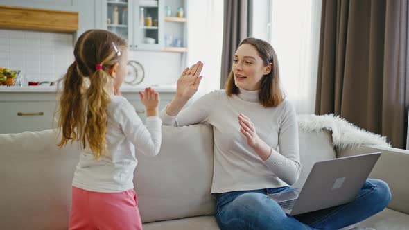 Young Mother Clapping Hands with Her Little Daughter Resting in Online Work on Laptop Home Interior