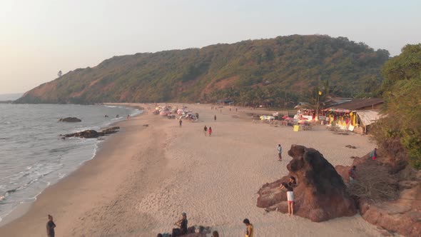 People stroll along Arambol beach, serene ocean waters. Goa, India. Aerial forward