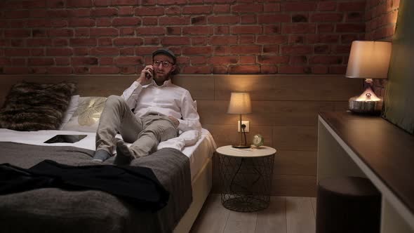 A handsome young businessman in a formal white shirt sits on a bed with a phone in a modern room.