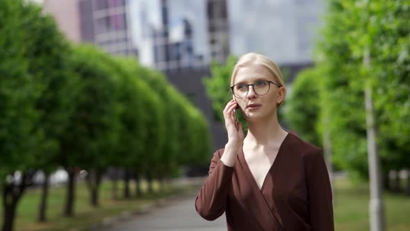 A Serious Confident Woman Answers the Phone As She Walks Down an Alley with Green Trees