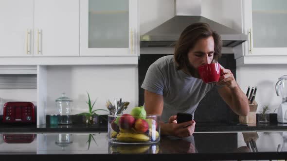 Caucasian man drinking coffee and using smartphone in the kitchen at home