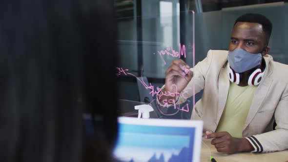 African american man wearing face mask writing on glass board at modern office