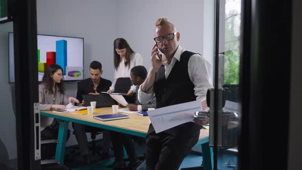 Portrait of Serious Confident Man Talking on Phone Analyzing Graph with Blurred Colleagues at