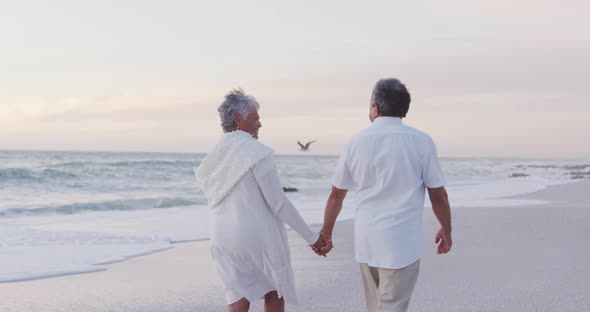 Back view of happy hispanic just married senior couple walking on beach at sunset