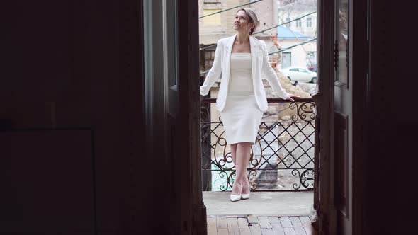 a Blonde Woman in a White Suit on the Balcony Against the Background of the City