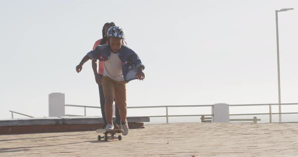 Video of happy african american father learning son how to skateboard on promenade