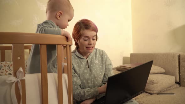 Young Mother is Working at Home Using Laptop and Taking Care of Her Little Baby Standing in the Crib