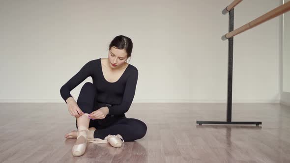 Ballerina in Black Tight Clothes Sits on the Floor and Puts on Pointe Shoes Next to the Ballet Barre