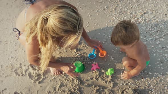 Mom with Glasses Plays with a Little Toddler Sitting on the Beach Near the Sea