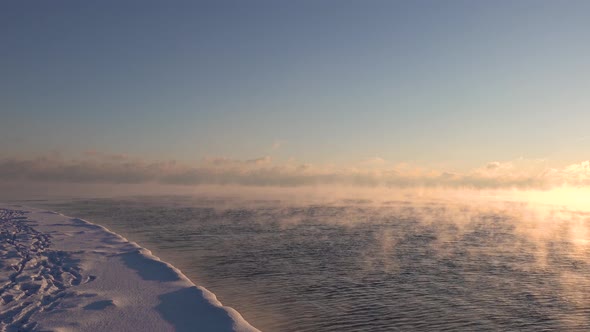 steam rising from a very cold lake as the morning sun rays shine on the water next to a snow covered