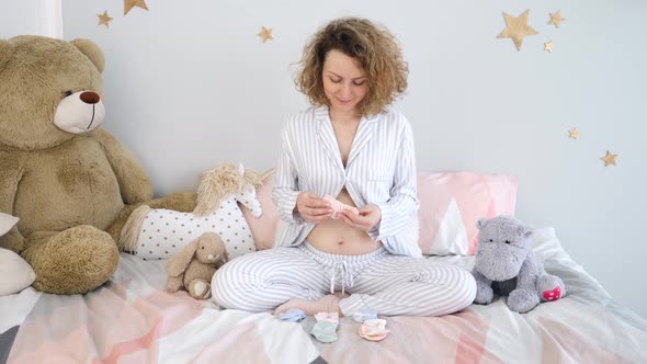 Young Smiling Pregnant Woman Choosing Socks For Future Baby Wearing Pajamas At Home.