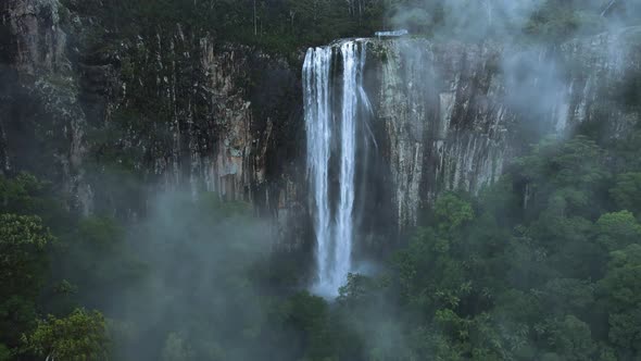 Unique drone view through suspended mist revealing a majestic waterfall spilling down a lush rainfor