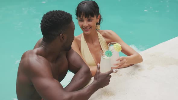 Happy diverse couple wearing swimming suits and drinking drinks at swimming pool in garden