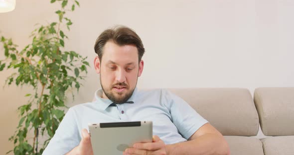 Young Bearded Man is Lying on the Sofa and Holding Tablet in Hands He is Relaxing and Scrolling the