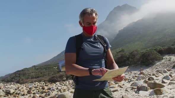 Senior hiker man wearing face masks with backpack reading map while hiking on the beach.