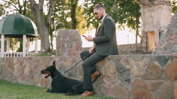 Wealthy Businessman in a Business Suit Is Using Smartphone and Smiling with a Doberman Dog