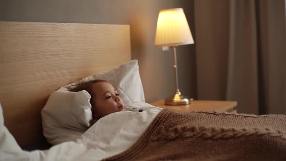 Adorable Little Sick Girl Lying in Bed Measuring Temperature with Thermometer in Mouth at Home