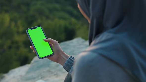 Woman in Hijab on Top of the Hill in the Evening Uses Smartphone with Green Screen on It