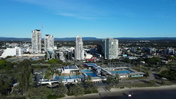 High panning view of a large Aquatic Centre on a coastal waterway with a towering urban city skyline