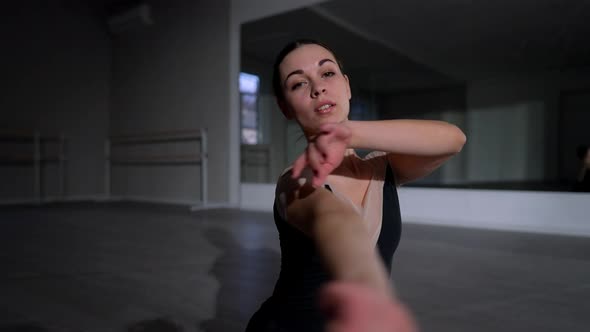 Slim Young Woman Looking at Camera Moving Hands Sitting on Floor in Dance Studio in Darkness