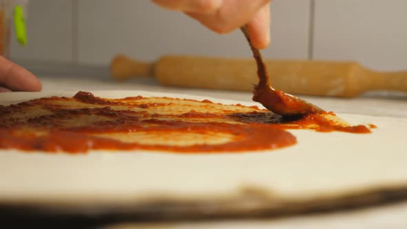 Male Arm of Cook Applying Ketchup on Pizza Dough Using a Spoon at Kitchen Table