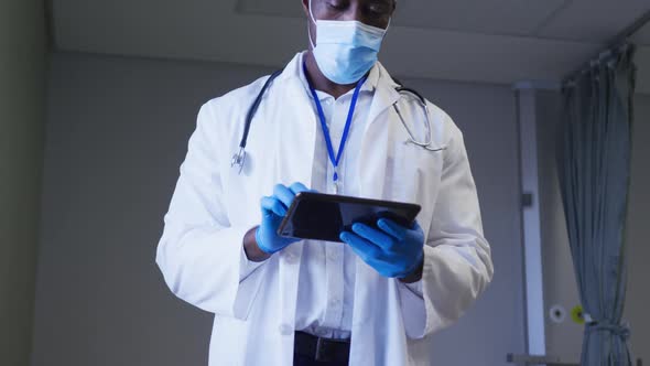 African american male doctor wearing face mask using digital tablet in hospital patient room