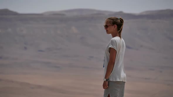 Young Woman in Sunglasses Standing on Cliff's Edge and Looking Around the Desert
