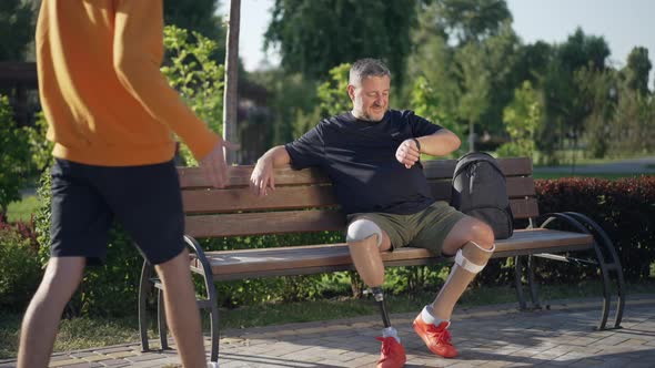 Handsome Cheerful Male Amputee Sitting on Bench in Park Checking Time on Watch Talking to Teenage