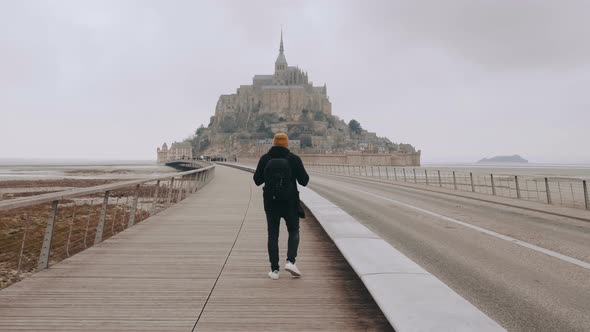 Camera Follows Handsome Happy Tourist Man Walking To Epic Mont Saint Michel Fortress, Smiling at