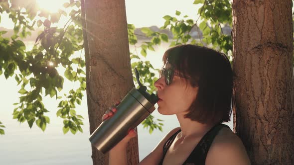 Sporty Woman Drinks From Metal Reusable Bottle Under Tree on the Riverbank