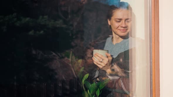 A Young Woman is Drinking Coffee From a Mug While Standing By the Window