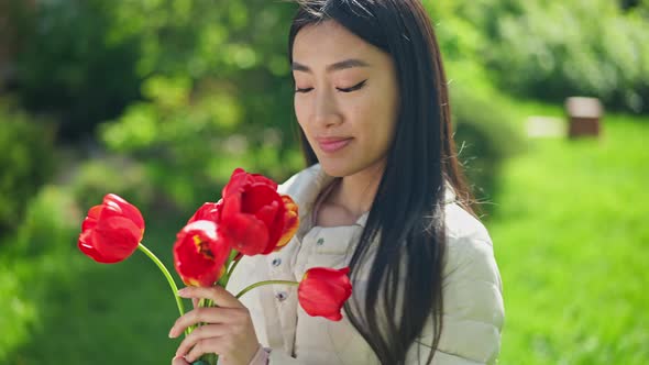 Slim Smiling Asian Woman Standing in Sunny Garden Admiring Bouquet of Red Tulips in Hand