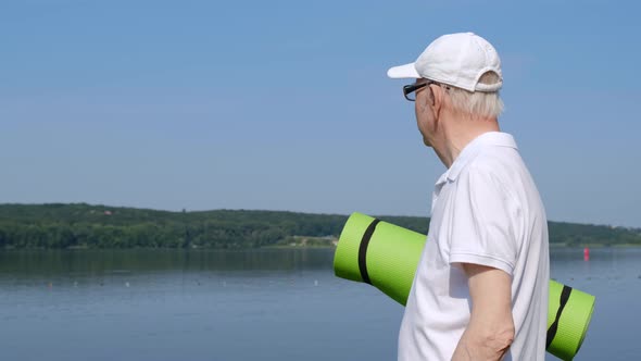 Grandfather retired in the morning city park, holding a yoga mat, looking at the lake.