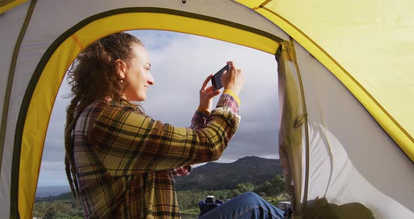 Happy caucasian woman camping, sitting outside tent on rural mountain taking photo with smartphone