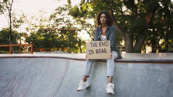 Afroamerican Woman Showing Cardboard Poster with Inscription the End is Near Posing Sitting on Pump