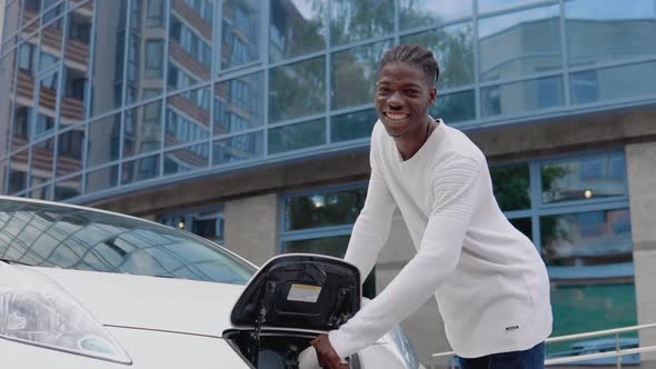 A Young African American Man Connects an Electric Car to the Charger and Looks at the Camera