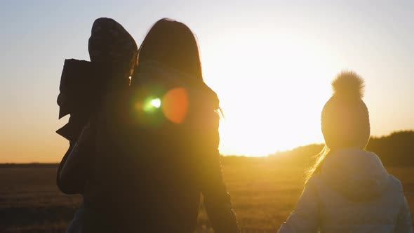 Happy Young Mother and Two Kids Walking on Meadow at Sunset. Silhouette of Friendly Family Concept.