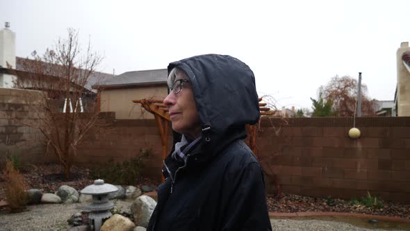 A middle aged woman who loves the rain wearing a raincoat hood and smiling as she watches the precip