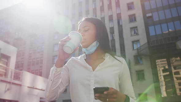 African american woman wearing face mask holding smartphone drinking coffee in city street