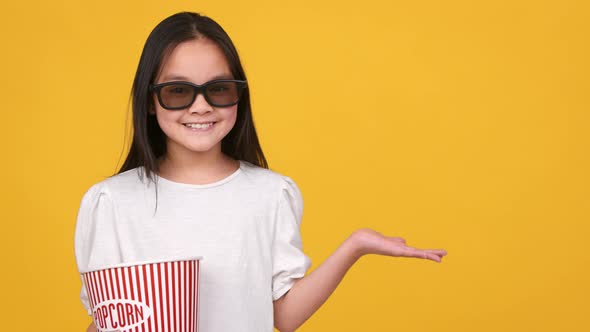 Happy Little Asian Girl in 3D Eyeglasses with Popcorn Bucket Showing Something with Outstretched