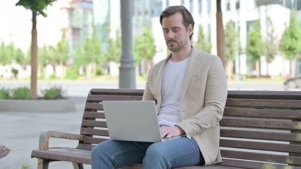 Young Man with Laptop Looking at Camera While Sitting on Bench