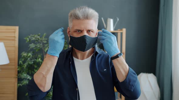 Portrait of Serious Guy Putting on Fabric Multiple-use Face Mask Looking at Camera in House alt