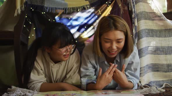 Asian mother reading a book and hugging her daughter under blanket fort at home