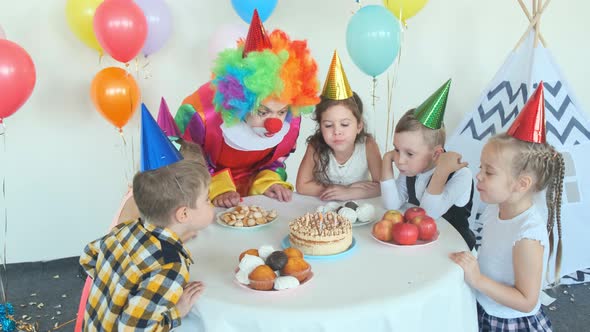 Little Kids and Clown in Wig Blow Candles on Birthday Cake