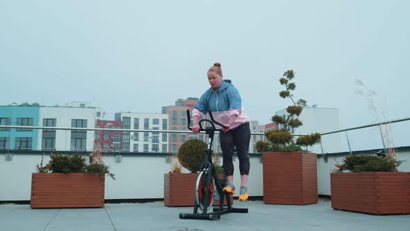 Healthy Caucasian Woman Exercising Workout on Stationary Cycling Machine Bike on House Rooftop Gym