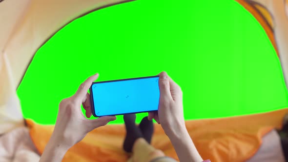 Man lying in tent uses smartphone with chromakey, against background of exit from the tent