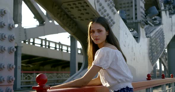 Lady in White Blouse Stands at Red Handrails Against Bridge