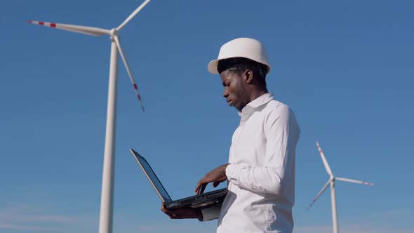 African American Electrician Engineer Standing on the Background of a Windmill at an Air Power Plant