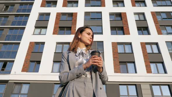 Young Woman in a Business Suit Drinks Coffee From a Plastic Cup on the Background of a Building on a